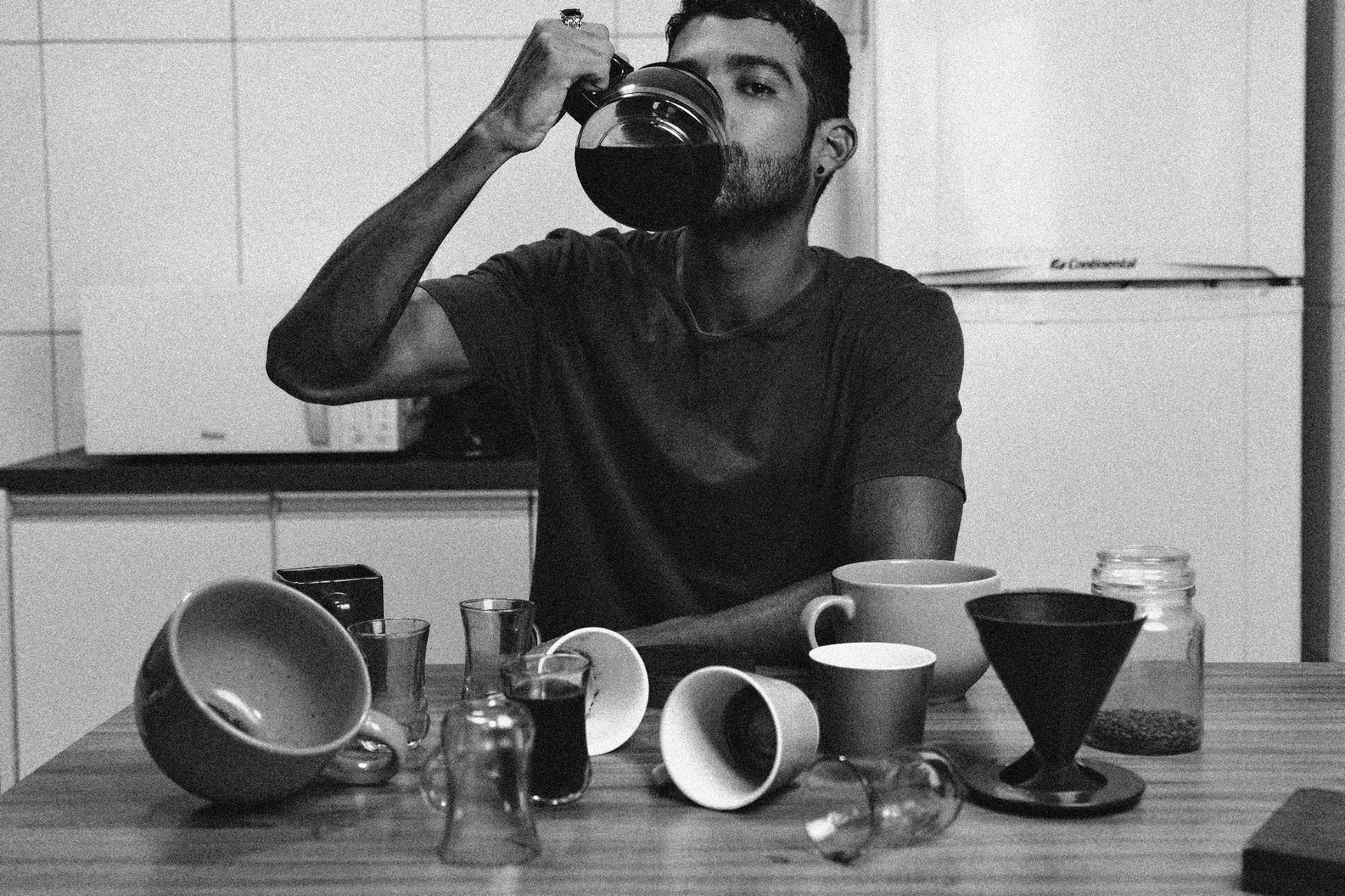 Tired night shift worker drinking coffee directly from a pot in a kitchen cluttered with cups, illustrating how caffeine becomes a coping mechanism